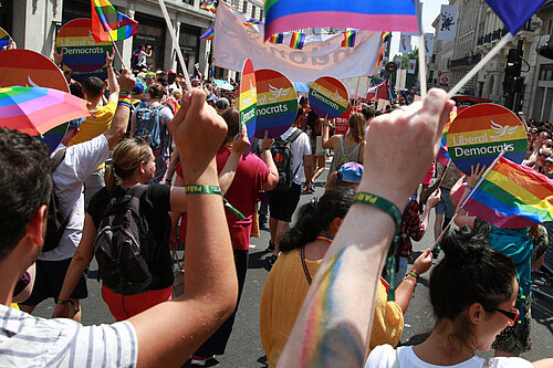 A Liberal Democrat group at Pride. The people are walking away from the camera, they are holding Lib Dem rainbow signs.