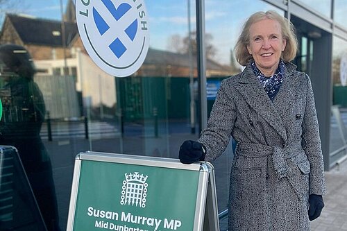 Susan Murray standing next to a sign advertising an advice surgery as MP for Mid Dunbartonshire.