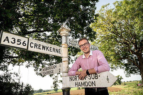 Adam Dance, MP for Yeovil, smiling and leaning against a vintage signpost at a rural crossroads. The signpost displays directions to Crewkerne, Ilchester, Martock, and North Perrott, set against a sunny countryside backdrop with trees and open fields.