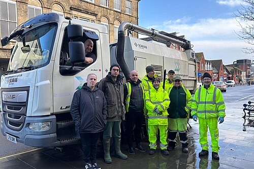 A group of workers in high-visibility yellow jackets stand together on a wet pavement next to a large street-cleaning vehicle with 'Somerset' branding. Adam Dance wears a dark jacket and green wellington boots and is standing among them, while a driver leans out of the truck’s window. The background features historic buildings and a town street with cars parked along the road.