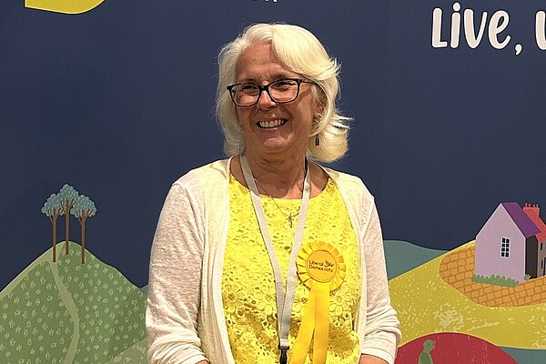 Sally Holland Stood in front of a Dorset Council Graphic banner. She is wearing a yellow dress and a white cardigan.