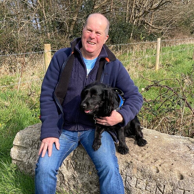 Nigel Amor sat on a log, with his black spaniel wearing a blue zipped jacket and jeans.