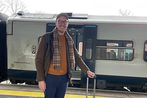 Adam Dance, MP for Yeovil, smiling on a train platform, holding a grey suitcase with a train in the background. He is dressed casually in a brown jacket, scarf, and cap, ready for travel.