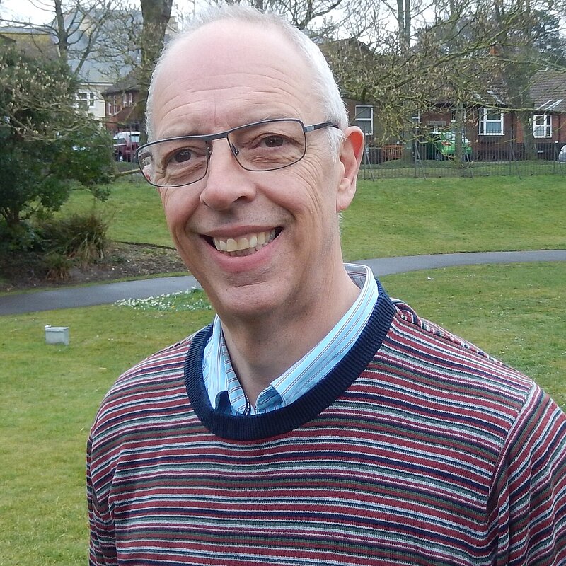 Gareth Jones is an older man with black glasses and wearing a jumper with red, white, blue, and green stripes & a light blue shirt