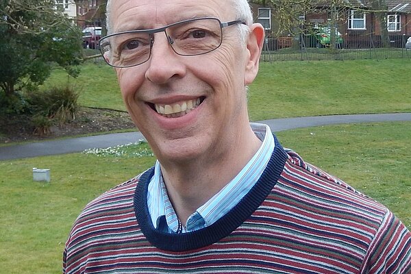 Gareth Jones is an older man with black glasses and wearing a jumper with red, white, blue, and green stripes & a light blue shirt