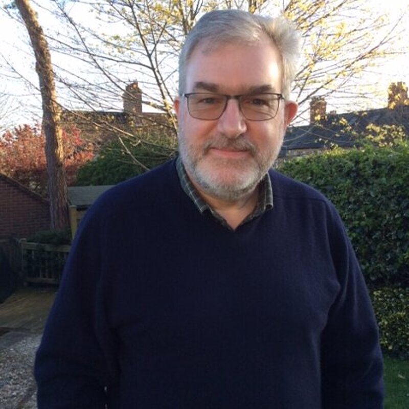 Paul Jacobs has grey hair and a beard. He is wearing a navy jumper and a green checkered shirt. He is in a garden
