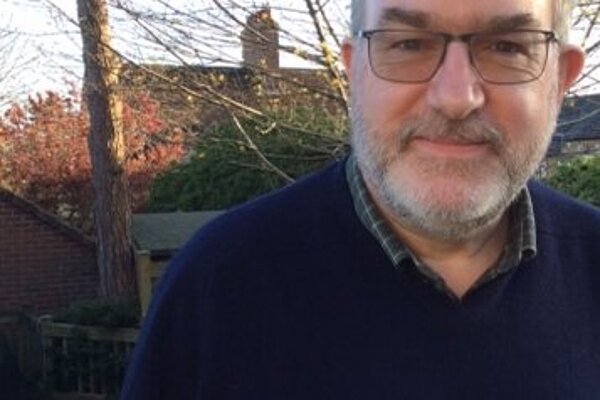 Paul Jacobs has grey hair and a beard. He is wearing a navy jumper and a green checkered shirt. He is in a garden