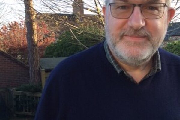 Paul Jacobs has grey hair and a beard. He is wearing a navy jumper and a green checkered shirt. He is in a garden