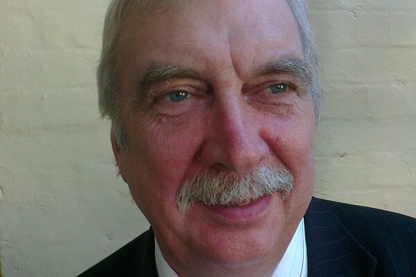 Jonathan Bourbon has grey hair and moustache. He is wearing a black suit with a white tie and white shirt