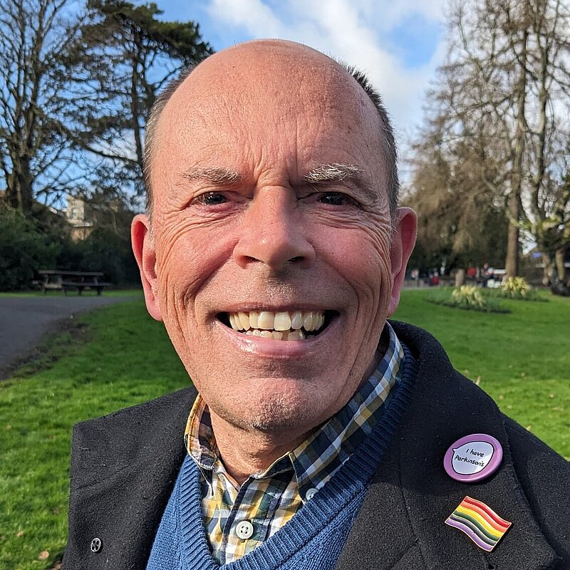 Andy Canning wearing a blue jumper, a yellow checked shirt and black suit jacket with a LGBTQ+ pride pin and a "I Have Parkinsons" pin.