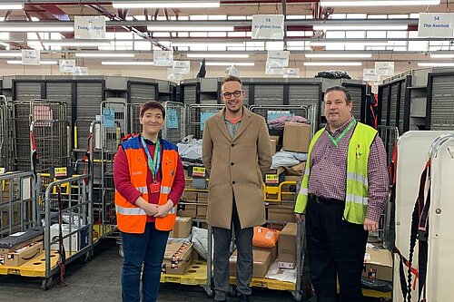 Adam Dance MP standing in a Royal Mail sorting facility alongside two staff members. He is wearing a beige coat and glasses, while the staff wear high-visibility vests, one orange and one yellow. They are surrounded by trolleys filled with parcels and letters, with visible signage and red beams in the background.